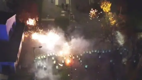 BBC Aerial image of fireworks igniting in a street at night in front of a line of police officers.