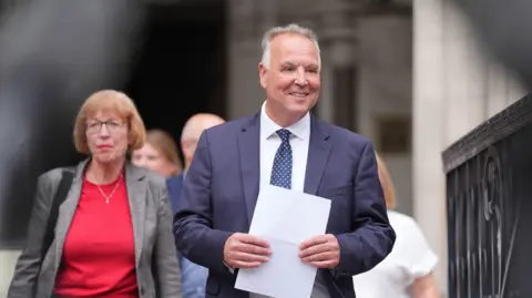 PA Media Chris Whitbread walking while smiling and clutching a piece of A4 paper. He has grey hair and is wearing a blue suit jacket over a white shirt, with a blue tie that includes white dots. To his right, slightly in the background, is a woman with brown hair and glasses and she is wearing a red top and grey blazer - she is the monitoring officer of Epping Forest District Council.