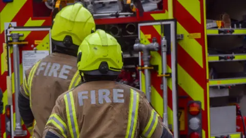Getty Images Back of three firefighters wearing cream uniforms saying 'fire' on the back. They are stood near a red fire engine. 