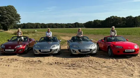 Glynis Jenkins Four Mazda MX5 are lined up next to each other on a green field. The two on the ends are red and the two in the middle are silver. Each car has a person stood in the middle of it looking towards the camera.  