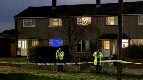 PA Media Two police officers in high-visibility clothing stand behind a cordon at night. There are three terrace houses behind them. Most of the windows are lit up by bright lights but some have curtains and blinds drawn.