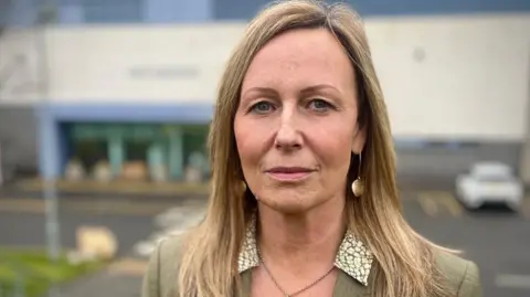BBC Head and shoulders shot of a woman with long blonde hair - combed in a middle parting - with a serious expression on her face. A prison building can be seen blurred in the background. 