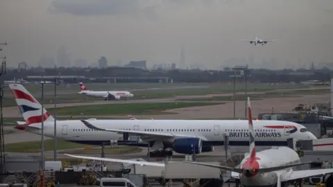 Reuters Planes arriving and departing from the runway at London Heathrow. One plane is coming into land, whilst others traverse the runways on the ground, where a BA plane parks at a gate whilst another smaller Swiss Airline plane also taxis behind it. The London skyline is clearly visible in the distance on a grey cloudy day, with the Shard being especially prominent. 