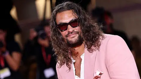 Maria Moratti/Getty Images Jason Momoa attending the Cartier Glory To The Filmmaker Award 2025 and the "In the Hand of Dante" red carpet during the 82nd Venice International Film Festival. He has long, wavy dark brown hair and a dark beard. He is wearing a pink suit jacket over a white shirt which is partially unbuttoned and black rimmed sunglasses. The blurred image of photographers can be seen behind him.