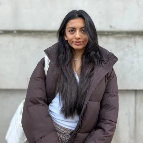 BBC A woman wearing a dark brown padded coat with a white shoulder bag visible on the left side, standing against a light grey concrete wall. Long dark hair falls over the coat, and a thin silver necklace is visible at the neckline.