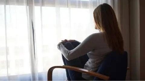 Getty Images A woman sits in a chair looking out a window, through curtains. You can just see the side of her and back of her head