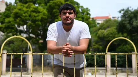 NICOLAS TUCAT/AFP via Getty Images Amine Kessaci is leaning on a yellow fence and looking straight at the camera. He has a white t-shirt on, and has short dark hair, and a slight beard.
