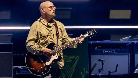 Getty Images Paul 'Bonehead' Arthurs, wearing glasses and a khaki hooded top, plays a brown-toned guitar in front of  a grey and black audio monitors on stage