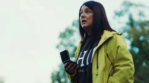 BBC A woman confronts Mark Daly in the park, holding a mobile phone in her right hand. She has long brown hair and is wearing a yellow jacket and a dark grey jumper.