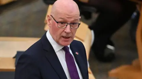 Getty Images John Swinney, who is bald and wearing glasses, speaks in the Scottish Parliament with his right hand held in front of his body, with the palm facing towards him. He is wearing a dark suit, white shirt and purple tie. 