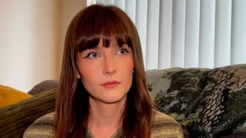 Abbie has long, wavy brown hair and is sitting on a sofa in her living room. She is is wearing a striped sweater in shades of brown and gray. Behind her are cushions including a green one, and a throw with a detailed map-like design. A white vertical blind covers the window in the background.