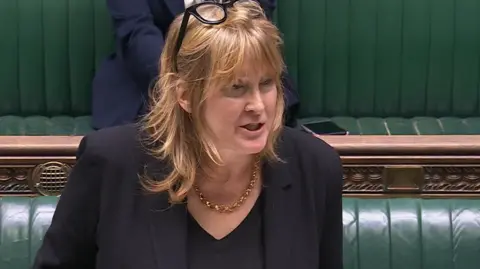 UK Parliament A woman with long fair hair stands at the Commons despatch box with green benches behind her. She is wearing a black suit and top, and a gold necklace.