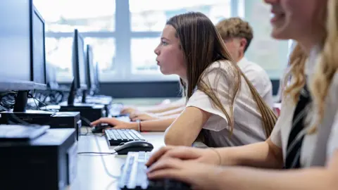 Getty Images Profile of a teenage girl with long hair in school uniform in a classroom looking closely at a computer screen. Fellow students sit either side of her.