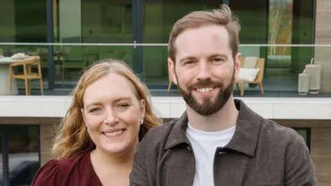 Omaze A man and woman both with blonde hair smiling at the camera while stood in front of a large house