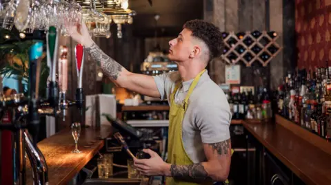 Getty Images A bartender wearing a grey polo shirt with a yellow apron on top hangs up glasses after washing them. 