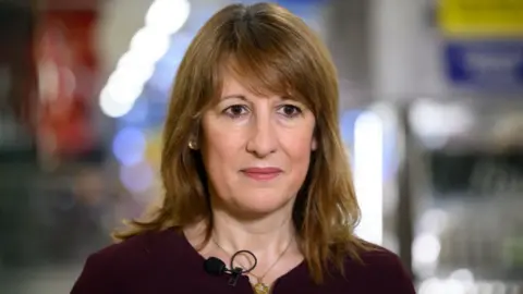 Getty Images Rachel Reeves, who has shoulder-length, brown hair, looks towards the camera in a close-up shot. She is wearing a burgundy top with a small black microphone clipped to the collar. 