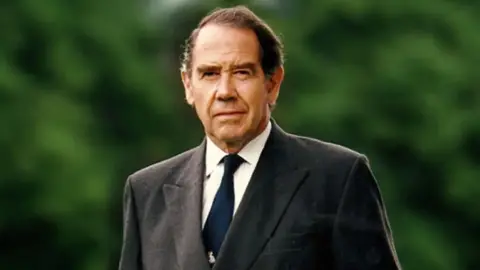 Sir Terence English Sir Terence walks towards the camera looking at the lens, with one eyebrow slightly raised. We can just see him from the chest upwards. He wears a grey suit jacket, white shirt and blue tie. He has short dark hair and is clean shaven.