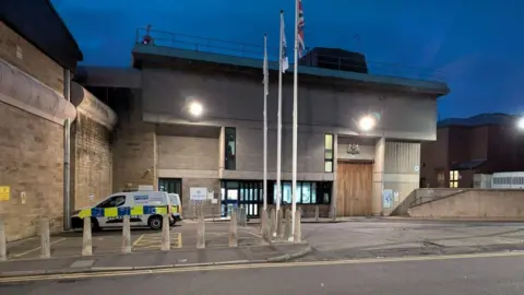 BBC Exterior of HMP Wakefield at night, with flags and police vehicles outside.