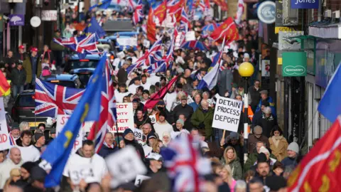 Eddie Mitchell Hundreds of people marching on a town high street. Some people are carrying union jacks and Sussex county flags, while one person holds a sign that reads "detain process deport".
