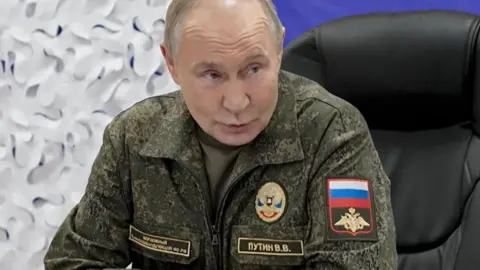 EPA Russian President Vladimir Putin speaks during a briefing to journalists at a military base. He is wearing a camouflage uniform and sat in a black office chair. The background of the image is a blue and white backdrop.