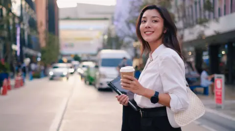 Getty Images A young woman with tablet and coffee in-hand crosses a street in a city centre. She is dressed in office attire, a white blouse and dark trousers.
