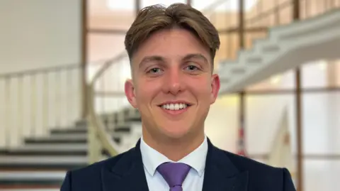 BBC/Simon Gilbert A man with brown hair, cropped at the sides, smiles at the camera in front of a curved staircase in a council building. He is wearing a black suit, white shirt and purple tie. 