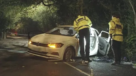 BBC Two police officers in yellow high-vis jackets inspect a white car that is crashed at the side of the road. 