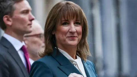 Reuters Rachel Reeves, who has brown shoulder-length hair, stands on Downing Street turned to her right, towards the camera. She is wearing a blue jacket and white scarf. 