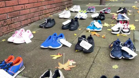 BBC More than 15 pairs of shoes, with labels attached, laid out in lines on the pavement