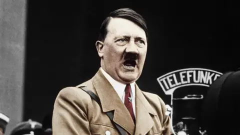 Getty Images A colour treated photo of Hitler speaking into a big microphone. He's wearing a light brown jacket and red tie.