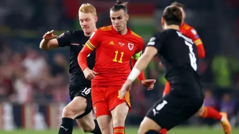 Getty Images Gareth bale in Wales kit dribbling between two austrian players in black kit. Gareth has an intent look and has his usual Bale bun ton knot hairstyle 