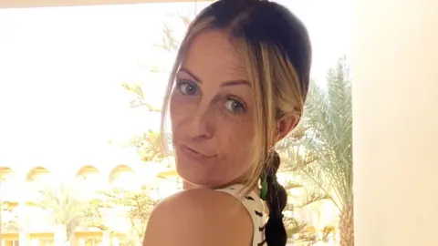 BBC Kirsty Buchan looks over her shoulder. She has blonde and brown hair tied in a plait and is wearing a white and black striped vest top.