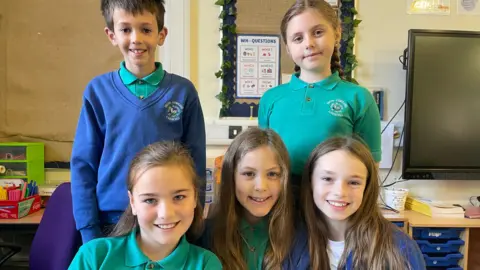 BBC Five pupils wearing green polo shirts in two rows in a classroom with two pupils standing at the back and three girls sitting in front of them. There is one boy standing, wearing a  blue jumper. All the children are smiling. 