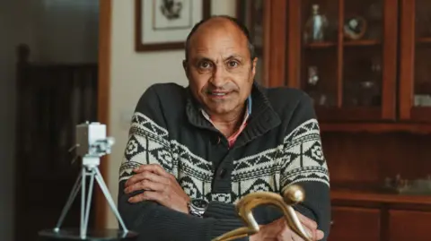 England Hockey A man with short hair, wearing a grey and white patterned top, his arms folded. He is smiling at the camera