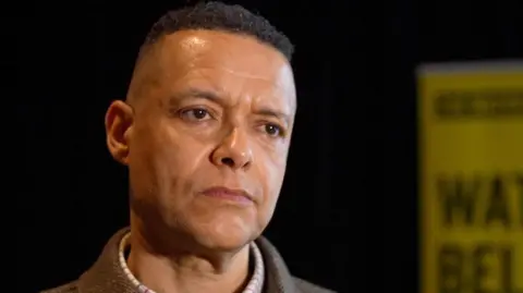 Martin Giles/BBC Cliver Lewis is left of the shot, standing inside and wearing a checked shirt, jumper and tweed jacket. He has short dark hair and there is a yellow sign in the background.