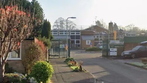 Google The outside of Friars Academy, showing a school, its gates, a car to the right, signs, railings, trees to the right and a lamp post.