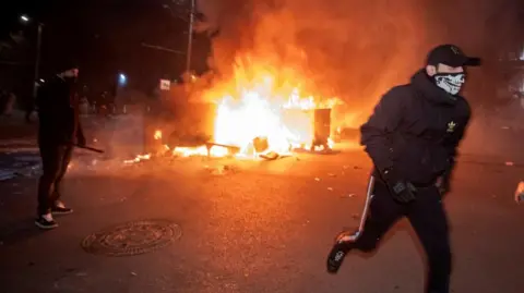 Reuters A masked protester in a baseball cap runs away from two flaming bins, with a line of shield-carrying riot police in the background.