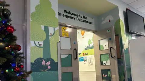 An interior shot of a children's hospital. A doorway to the emergency department is seen sitting open. The walls are painted with cartoon like images of nature. A Christmas tree can be seen to the left