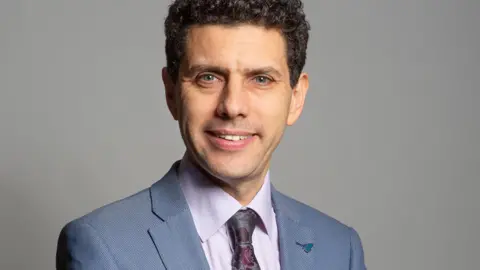 UK Parliament A man with dark hair and blue eyes looking at the camera. He is wearing a blue suit jacket, a lilac shirt and a pink and grey tie. There is a blue pin on his lapel. He is stood in front of a grey background.