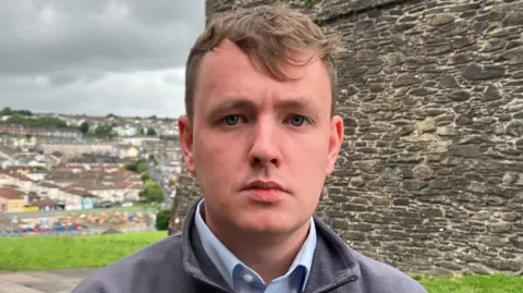 Sinn Féin assembly member Pádraig Delargy is in a grey fleece and blue shirt. He is standing beside Derry's historic walls. A number of homes can be seen in the background.