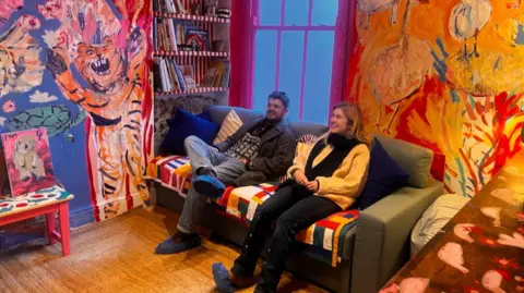 A couple sit back on a sofa and look at the circus animals and big top painted on the walls