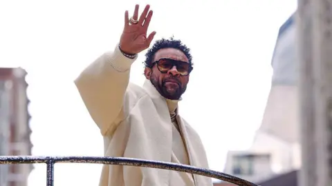 Getty Images Shaggy wears a large, off-white woolen coat as he waves at a crowd below. He is standing on a raised platform or balcony, with tall buildings just visible in the background.