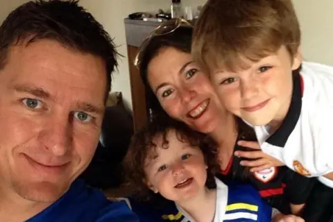 Anna-Louise Bates A man and woman are smiling. The woman has a young girl on her knee and a little boy sitting next to her. They are in a living room, with a fireplace and a wall-mounted TV seen behind them.