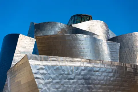 Tim Graham/Getty Images A focused photograph of the Guggenheim Museum in Bilbao. The building's iconic titanium cladding shines in bright sunlight with a glass box sticking out at the top building