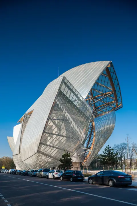 Michael Jacobs/Art in All of Us/Corbis via Getty Images The Louis Vuitton Foundation building is seen against a bright-blue sky. Its steel and glass structure bursts out of the ground in a variety of wings surrounding a central brick building that is obscured from view by the glass. It sits beside parked cars on a road with trees in the background.