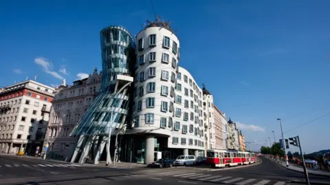 Insights/Universal Images Group via Getty Images Prague's Dancing House is seen in a portrait photograph taken from the street. A tram passes by near traffic. The building's unique slanting frontage is seen covered in a concrete and glass concoction. Gehry's iconic window-style is also seen on the building's facade.