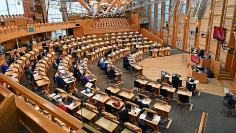 Getty Images Image of the Scottish Parliament chamber while MSPs are voting on amendments to a bill - the room is a large semi-circular arena with individual wooden desks for each member, and paperwork and laptops are much in evidence