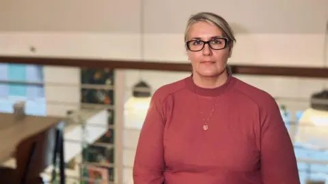 Kay Davies, who has blonde hair and is wearing glasses and a pink top, is sitting on a bench in a café