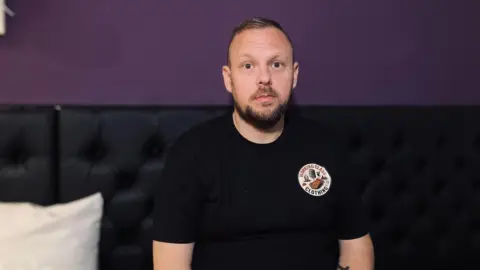 Kevin Robinson-Hale, who has a short beard and is wearing a black t-shirt, is sitting on the mezzanine level of a cafe