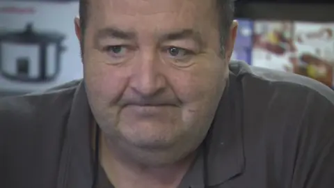 Gerard Woodhouse, in a close-up shot, looking away from the camera. He is wearing a dark grey shirt. 